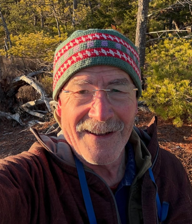 Smiling person in a colorful knit hat, standing in a forest with trees and sunlight in the background.