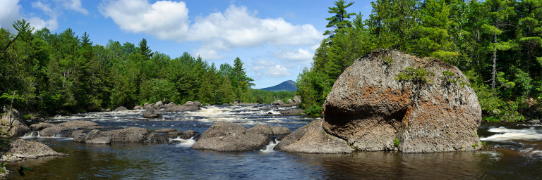 A serene river flows through a forested landscape, surrounded by large rocks and distant mountains under a blue sky.