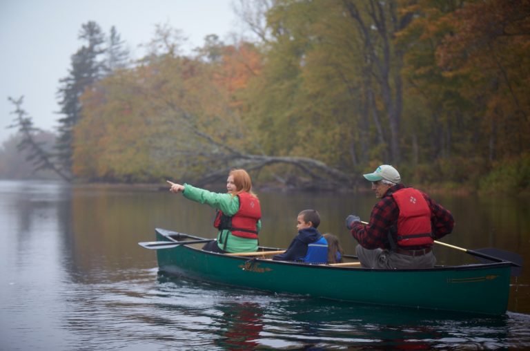A woman points out something while canoeing with two children on a serene, foggy lake surrounded by autumn foliage.