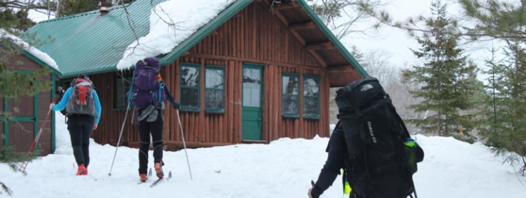 Three hikers approach a wooden cabin covered in snow, surrounded by pine trees. Winter scene with clear skies.