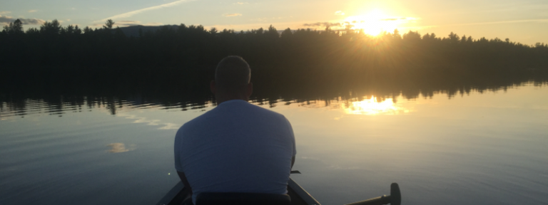 A person sits in a canoe, gazing at a sunset reflected on still water, surrounded by trees.