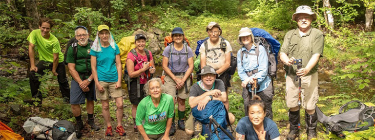 A diverse group of hikers poses together in a lush green forest, all wearing backpacks and outdoor gear.