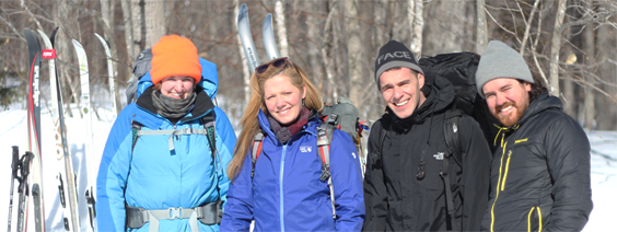 Four people smile for a photo in a snowy landscape, with skis and trees in the background.