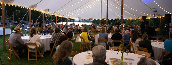 A large outdoor event under a festoon-lit tent, with many attendees seated at round tables, listening to a speaker.