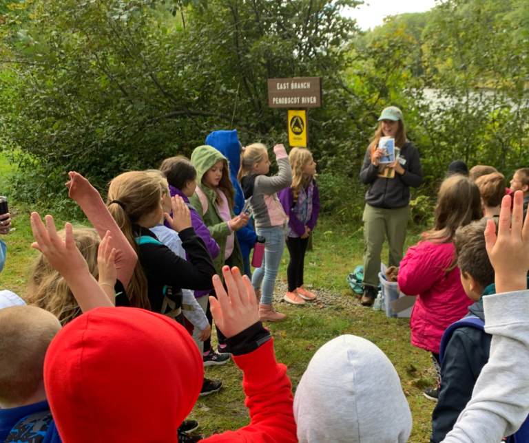 A group of children raising hands while a guide shares information near the East Branch of the Penobscot River.