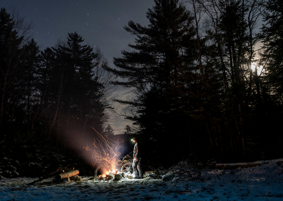 A person stands by a campfire in a snowy forest, stars twinkling above and a bright moon illuminating the scene.
