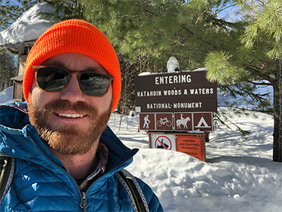 Smiling person in a blue jacket and orange beanie stands by a snowy sign for Katahdin Woods & Waters National Monument.
