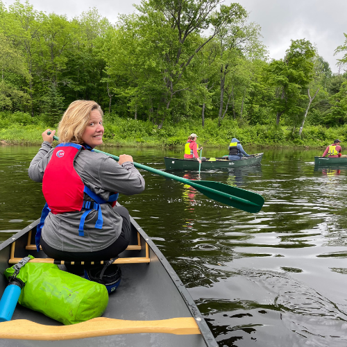 A woman in a canoe smiles at the camera, paddling in a serene river with others in the background. Green trees surround the scene.
