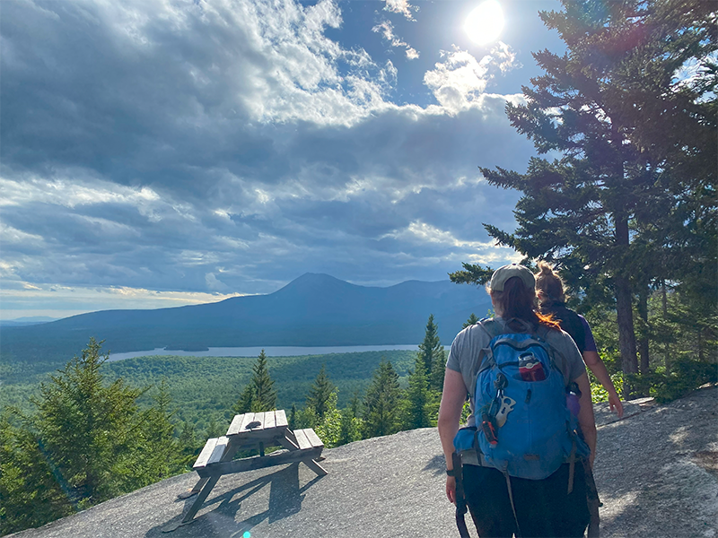 Two hikers walk towards a scenic overlook with mountains and a lake under a partly cloudy sky.