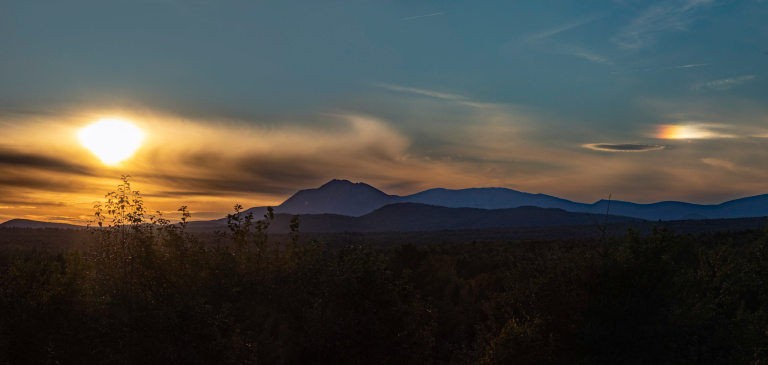 Sunset over mountains with vibrant orange and blue skies, silhouetted trees in the foreground.