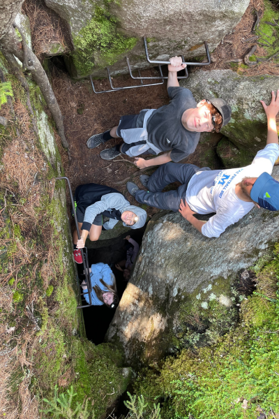 Four young teens smile up at the camera from a cave entry with iron climbing rungs in a mossy rock.