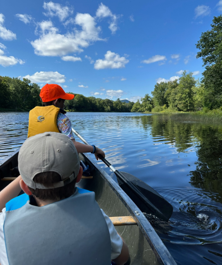 Two children canoeing on a calm lake surrounded by lush greenery under a bright blue sky with fluffy clouds.