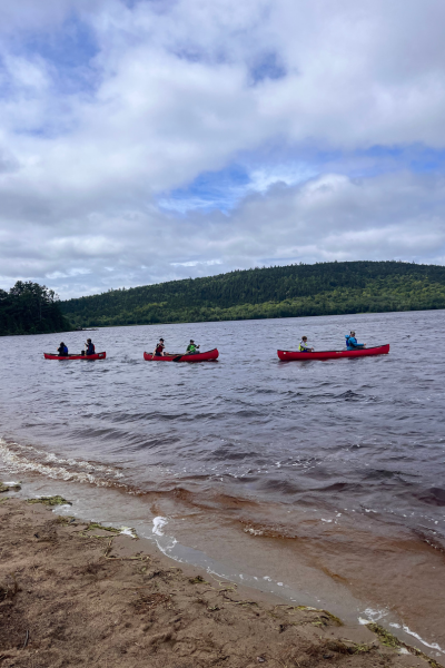 A line of six red canoes on a lake, with a backdrop of green hills and cloudy skies.