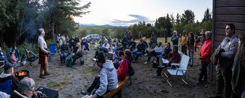 A group of poeople sitting in camp chairs around a campfire and woman speaking. 