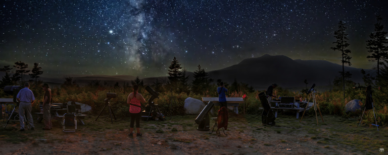 A group of people observe the starry night sky using telescopes, with mountains and trees in the background.