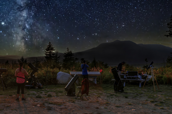People observe the night sky with telescopes amidst trees and mountains under a starry sky.