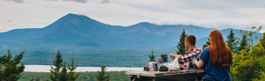 Two people seated at a picnic table facing a dramatic mountain behind a lake.