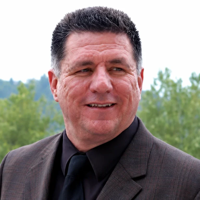 A man in a suit smiles outdoors, surrounded by greenery and soft hills in the background.