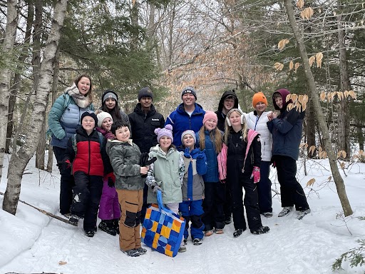 A group of 15 people, including children and adults, pose together on a snowy path in a forest.