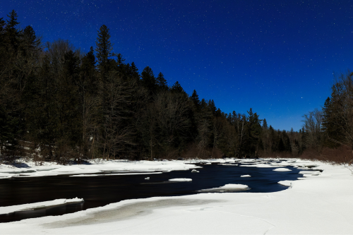 A serene winter landscape featuring a dark river flowing through a snowy bank, surrounded by trees under a starry night sky.