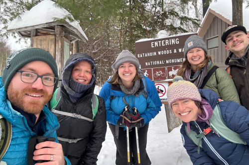 A group of six friends poses smiling in snow near a sign for Katahdin Woods and Waters National Monument.