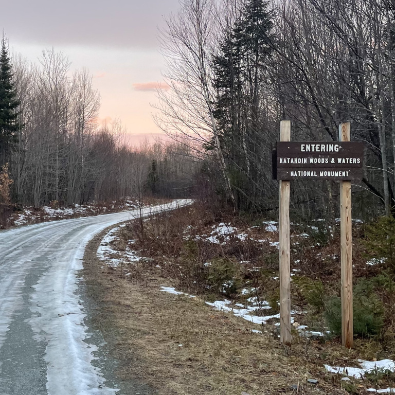 Sign for "Entering Katahdin Woods & Waters National Monument" beside a winding, snowy road lined with trees at dusk.