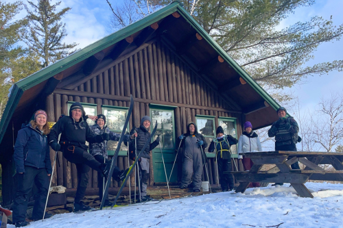 A group of eight people stands outside a cabin in the snow, smiling and holding ski poles, with trees in the background.