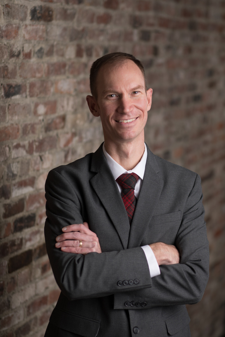 A man in a gray suit and patterned tie smiles confidently with arms crossed against a brick wall backdrop.