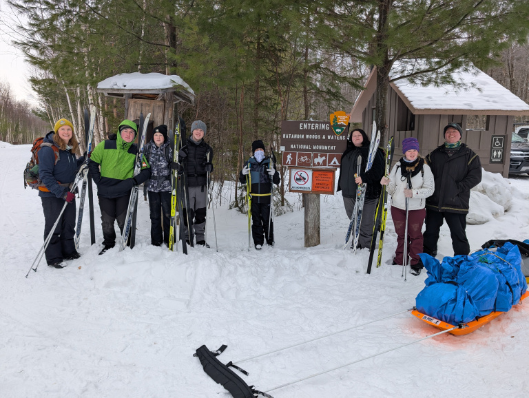 A group of seven people in winter gear stands near a snowy trailhead sign, holding skis and a sled.