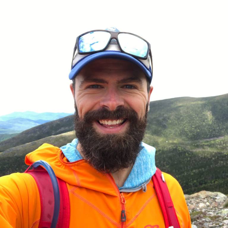 A smiling man with a beard wearing an orange jacket and a blue cap, stands atop a mountain with a scenic backdrop.