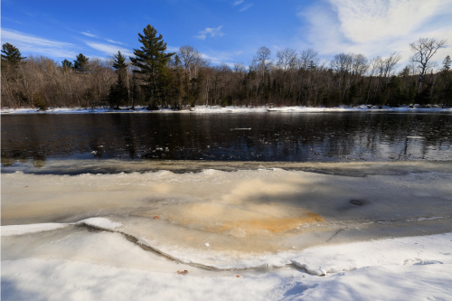 Winter scene of a partially frozen river, with blue sky and trees lining the bank.