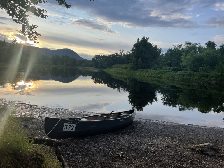 A canoe rests on a pebbled shore beside a calm river reflecting the sunset and surrounding trees.