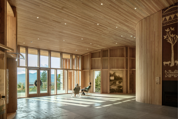 A spacious wooden interior with large windows, natural light, and two people relaxing in chairs.
