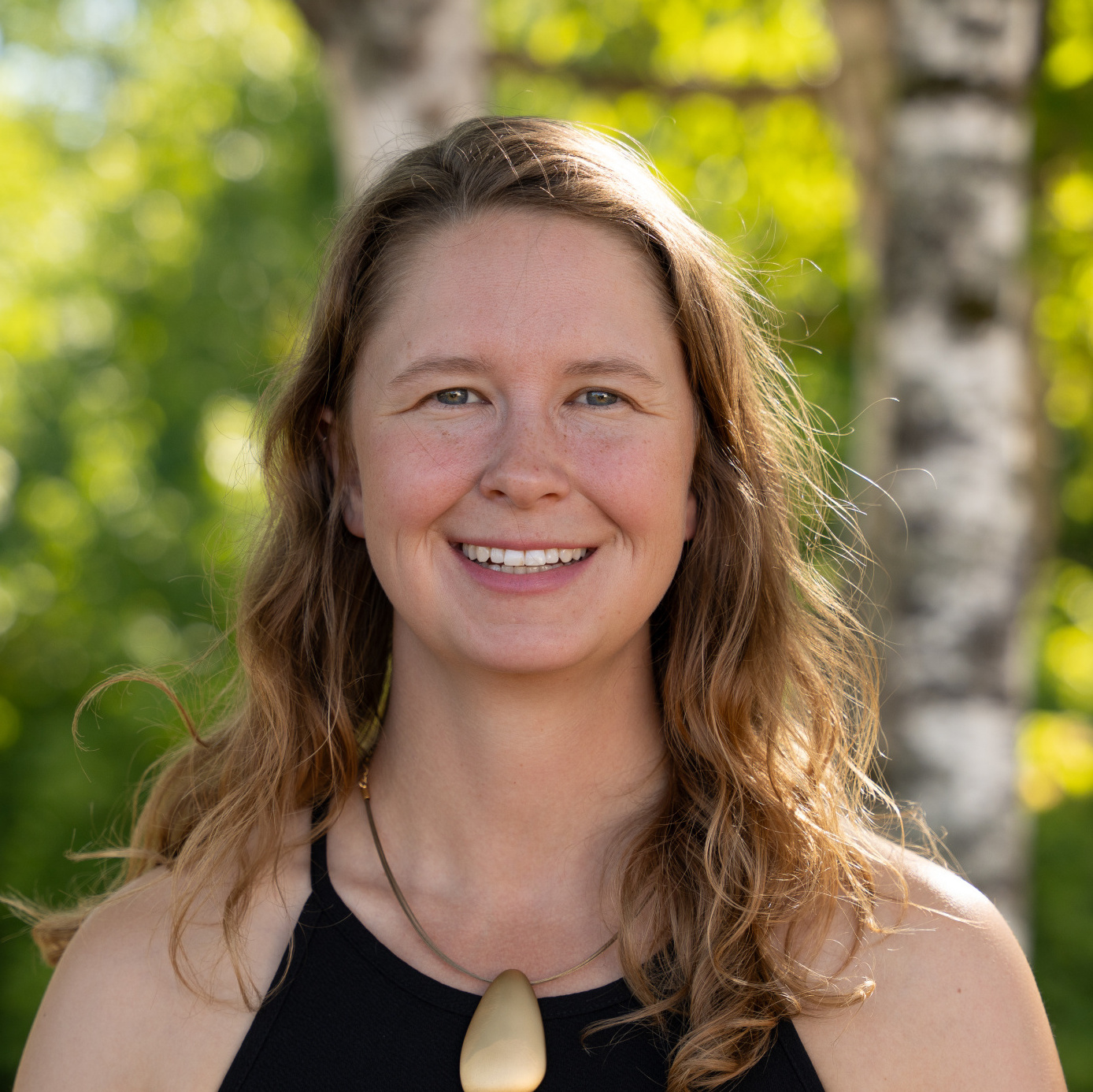 Smiling woman with long, wavy hair in a black top, set against a lush green background with birch trees.