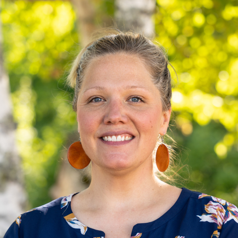 Smiling woman with light hair, wearing a floral blouse and large earrings, stands against a green, leafy backdrop.