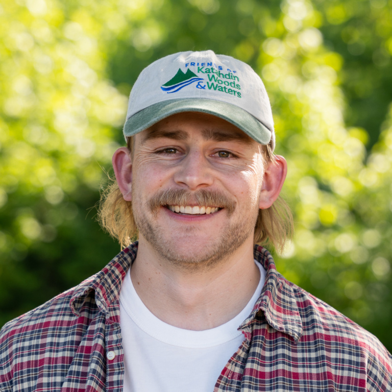A smiling man with a mustache wears a cap and plaid shirt, standing against a blurred green background.