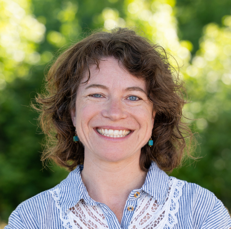 Smiling woman with curly hair wearing a striped shirt and earrings, set against a green, blurred background.