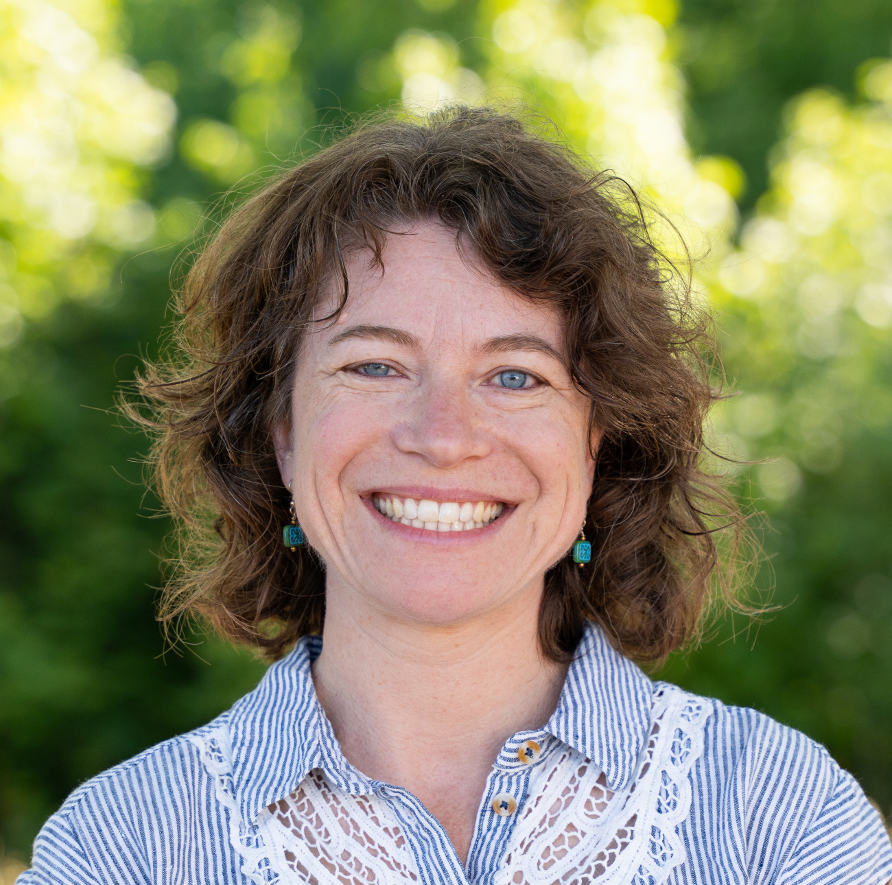 Smiling woman with curly hair wearing a striped shirt and earrings, set against a green, blurred background.
