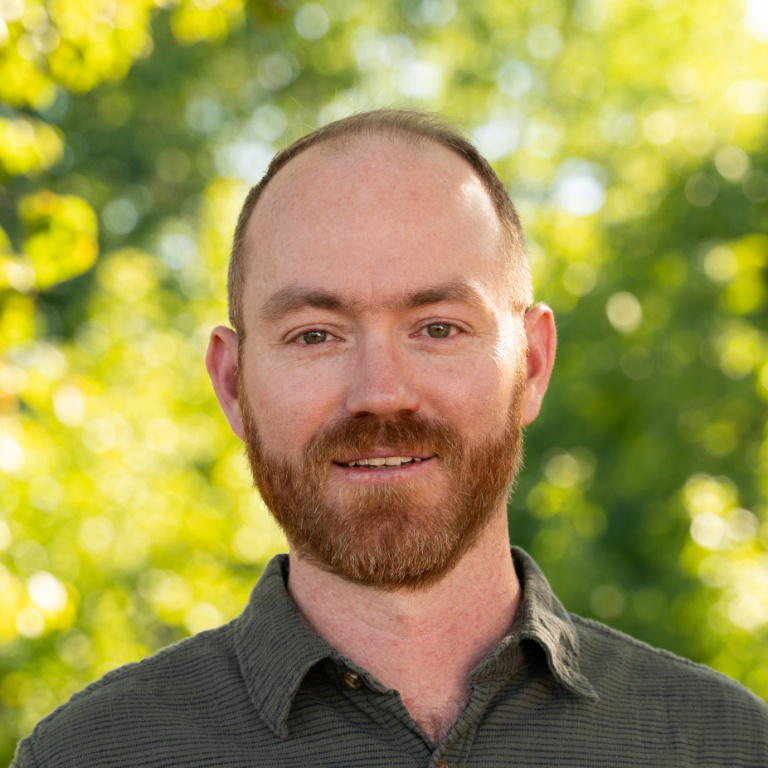 A smiling man with a beard stands outdoors, surrounded by soft, green foliage and dappled sunlight.