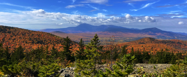 Vibrant autumn landscape with orange and green foliage, mountains, and a blue sky dotted with clouds.