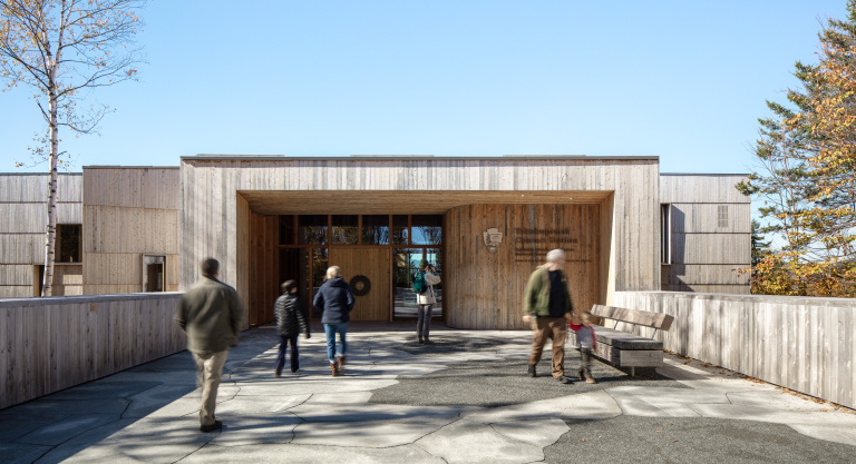 Wooden building entrance with people walking, surrounded by trees and a clear blue sky. A bench sits nearby.