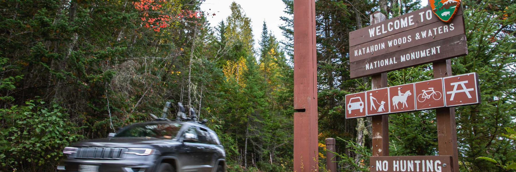 A car drives past a sign for Katahdin Woods and Waters NM with the NPS arrowhead logo.