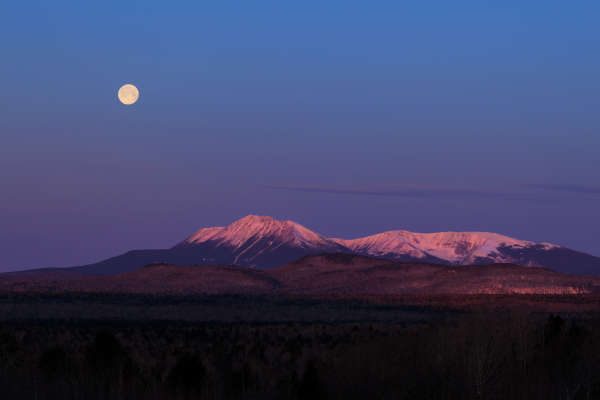 A serene view of snow-capped mountains at dusk, with a full moon glowing in a clear sky above.