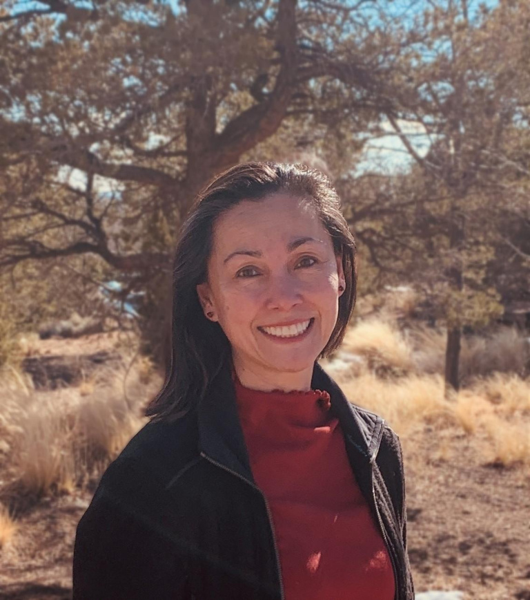 A woman smiles in a natural setting, with trees and dry grass in the background, wearing a red shirt and black jacket.