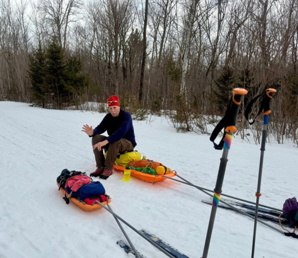 A person sits on snow-covered ground next to sleds, gesturing while surrounded by trees in a winter landscape.