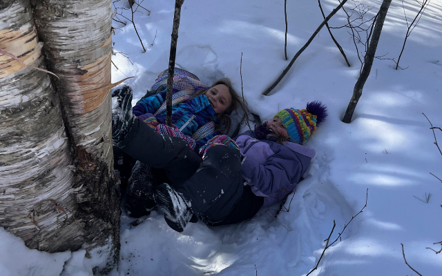 Two children play in the snow, lying together beside a tree, surrounded by winter scenery and sunlight.