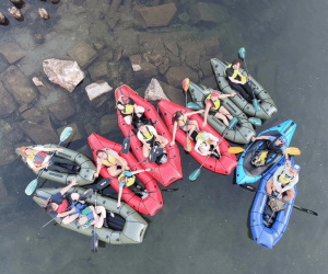 An aeirial view of nine inflatable rafts in a rocky-bottomed river's edge.