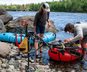Two white men attach a bicycle to the prow of an inflatable raft.
