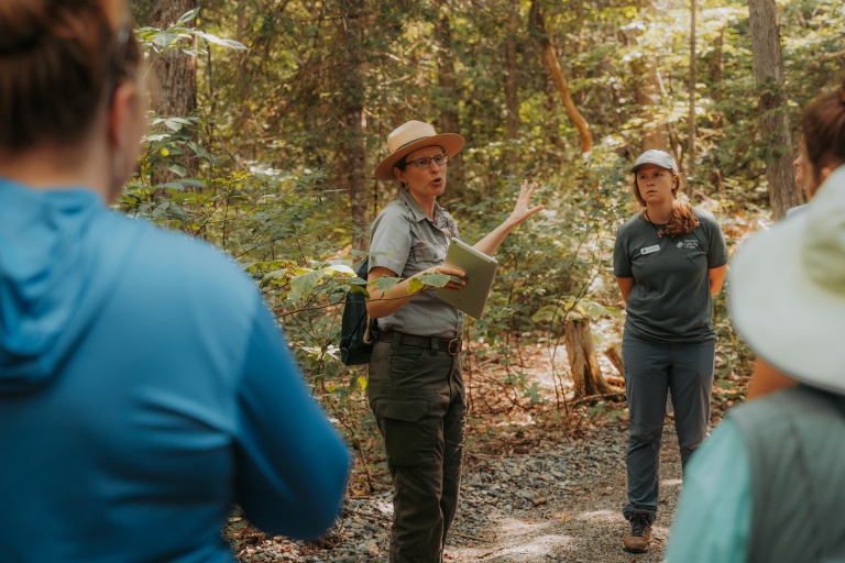 A park ranger explains nature to a group of visitors in a forest setting. Sunlight filters through the trees.