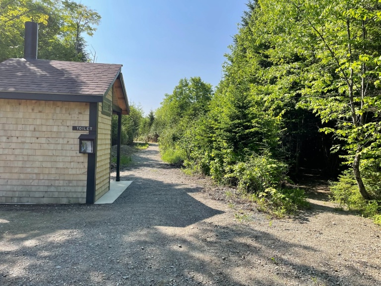 A rustic wooden bathroom building near a gravel path, surrounded by lush green trees and a clear blue sky.